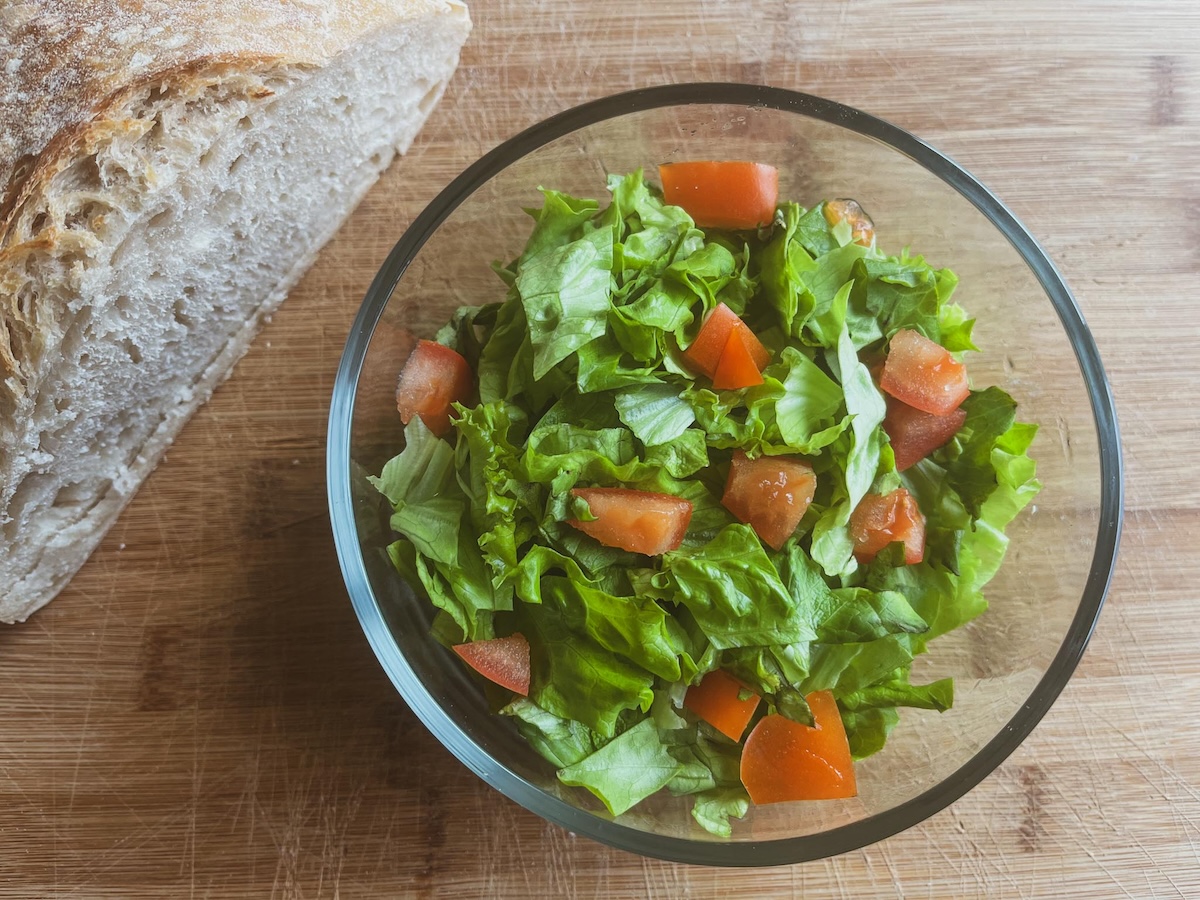 lettuce and tomato in a glass bowl with a load of sourdough on a wood surface